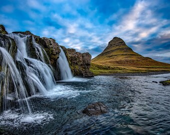 Op de afbeelding: Een waterval stroomt in een rivier voor een groene, grasbegroeide berg met een puntige top tegen een blauwe lucht met witte wolken.