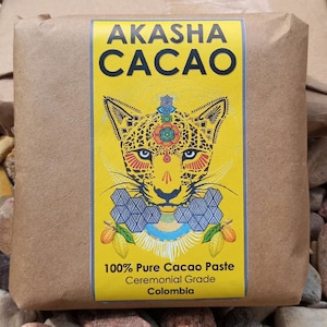May include: A brown paper-wrapped package with a yellow label featuring the text "AKASHA CACAO" and a colorful jaguar illustration. The label also states "100% Pure Cacao Paste, Ceremonial Grade, Colombia."