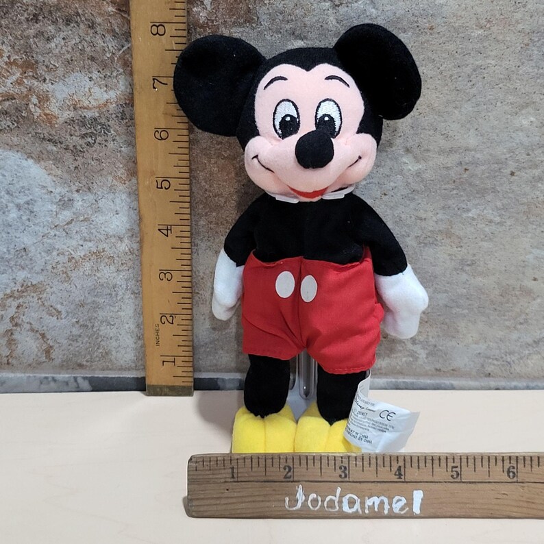 May include: A Mickey Mouse plush toy, featuring a black body, ears, and shorts, with red shorts and yellow shoes. The toy has white gloves and a smiling face. A ruler is next to the toy, measuring approximately 8 inches.
