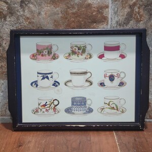 May include: A rectangular serving tray with a dark brown frame. The tray's surface features nine teacups and saucers with floral and patterned designs in various colors, including pink, blue, and green. The background is a light blue.