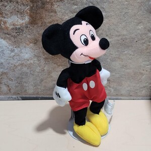 May include: A plush Mickey Mouse toy, featuring a black head and ears, a red shorts with white buttons, yellow shoes, and white gloves. The toy has a pink nose and a smiling expression. The toy is standing on a white stand.