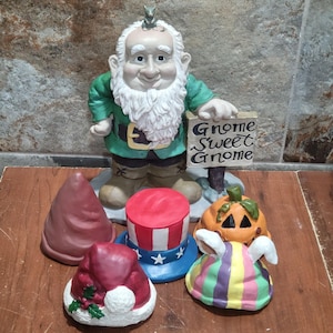 May include: A ceramic gnome figure with a white beard and a green jacket, holding a sign that reads "Gnome Sweet Gnome." Various decorative hats, including a Santa hat, a pumpkin, and a patriotic hat, are arranged around the gnome.