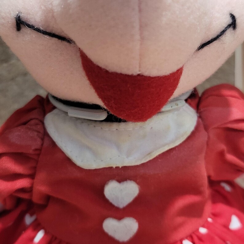 K&ouml;nnte beinhalten: Nahaufnahme des Gesichts und des Oberk&ouml;rpers einer Pl&uuml;schpuppe. Die Puppe hat ein rosa Gesicht, schwarz gen&auml;hte Augen und einen roten Mund. Sie tr&auml;gt ein rotes Kleid mit wei&szlig;en herzf&ouml;rmigen Akzenten und einem wei&szlig;en Kragen. Das Kleid der Puppe hat ein R&uuml;schendesign.