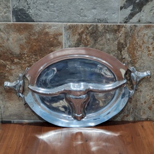 May include: A silver serving tray with a bull's head design. The tray has two handles on the sides and a raised rim.