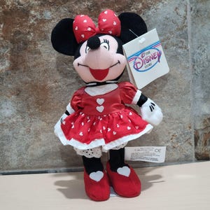 K&ouml;nnte beinhalten: Minnie Mouse Pl&uuml;schpuppe mit rotem Kleid mit wei&szlig;en Herzakzenten und passender Schleife. Die Puppe hat schwarze Ohren, ein l&auml;chelndes Gesicht und rote Schuhe mit wei&szlig;en Herzen. Ein Disney Store-Etikett ist angebracht.