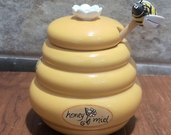 May include: A yellow ceramic honey pot shaped like a beehive with a white flower on top. The pot has a wooden honey dipper with a bee figurine on the end. The pot has the words "honey" and "miel" written on it.