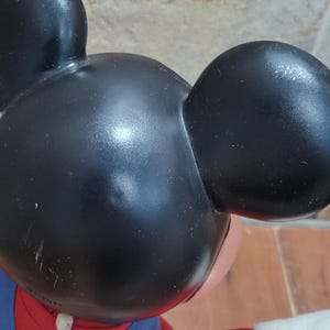 May include: Close-up of a Mickey Mouse figurine. The figure has a black head with large, rounded ears. The face is pink, and the figure is wearing a red and blue outfit. The figurine is a collectible item.