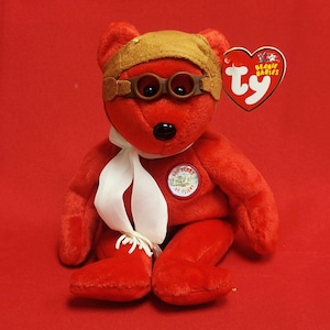 Puede incluir: Un oso de peluche rojo con gorro y gafas de aviador marrones, con una bufanda blanca y una etiqueta que dice "Beanie Babies". El oso tiene una nariz negra y un parche que dice "100 Years of Flight".