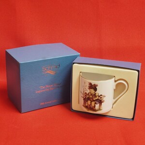 Può includere: Tazza in ceramica bianca con bordo dorato, con un'illustrazione infantile, presentata in una scatola blu. La scatola recita "The Flower Basket" e "1982 Annual Gallery". La tazza è inserita in un inserto color crema.