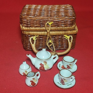 May include: A wicker picnic basket with a handle and a set of miniature porcelain teacups, teapot, and creamer. The teacups and teapot have a floral design with a red and white color scheme.