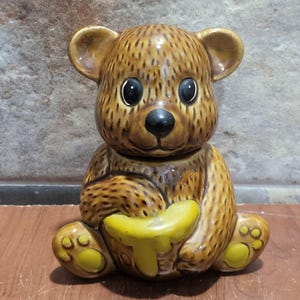May include: A brown ceramic bear-shaped honey pot with a yellow honey pot. The bear has large black eyes, a black nose, and yellow accents on its paws. The honey pot is sitting on a wooden surface.