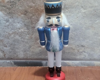 May include: A wooden nutcracker figurine in a blue uniform with a black hat and white beard. The figurine stands on a red base. The hat has a gold finial and blue and gold trim. The nutcracker's face is painted with a mustache and a stern expression.