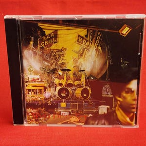 May include: A CD case featuring album art with a nighttime city scene. The artwork includes signs for a bar and arcade, a drum set, and a person in the foreground. The CD case is in a clear plastic jewel case.
