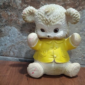 Peut inclure: Un ours en peluche blanc portant une veste jaune. L'ours a un nez brun et des yeux bruns. L'ours est assis sur une surface en bois.