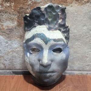 May include: A handcrafted ceramic mask with a human face. The mask features a textured surface with shades of gray, blue, and black. The eyes are hollowed out, and the mask has a rough, artistic finish, suggesting a unique, decorative piece.