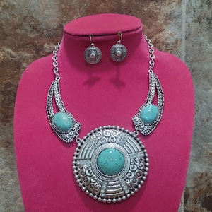 May include: Silver-toned necklace and earring set with turquoise-colored stones. The necklace features a large central pendant with intricate detailing and two smaller side pendants. The earrings are circular with matching designs.