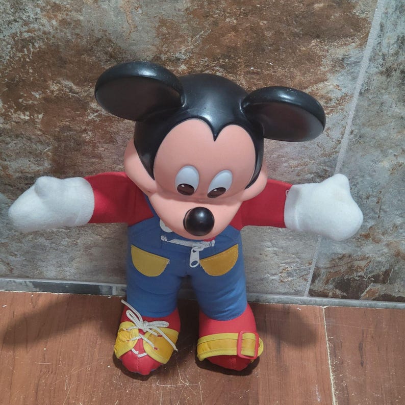 May include: A Mickey Mouse plush toy. The toy has a black head and ears, a peach-colored face, and a large black nose. It wears a red shirt, blue overalls with yellow accents, white gloves, and red shoes with yellow laces.