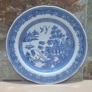 Peut inclure: Une assiette ronde en céramique blanche et bleue avec un bord festonné. L'assiette présente un motif Willow classique bleu et blanc avec un paysage pittoresque, comprenant un pont, des arbres et des oiseaux. L'assiette est une pièce décorative.