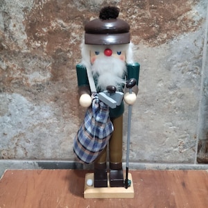 May include: A wooden nutcracker in the form of a golfer, wearing a brown hat and green jacket. The nutcracker has a white beard, a red nose, and is holding a golf club and a plaid bag. A golf ball is at the base.