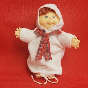 May include: A vintage doll with red hair, green eyes, and a smiling expression. The doll is wearing a white hooded outfit with a red and black plaid scarf. The doll's hands are outstretched, and it is standing on a red surface.