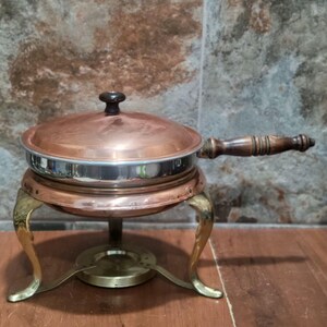 Peut inclure: Un plat à réchaud en cuivre avec un intérieur argenté, un couvercle en cuivre assorti doté d'un bouton sombre et une longue poignée en bois. Le plat repose sur un support en métal doré à trois pieds incurvés et un brûleur central.