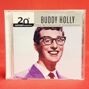 Peut inclure: Un boîtier de CD avec un portrait de Buddy Holly portant des lunettes à monture noire et une cravate rouge. Le texte "THE BEST OF BUDDY HOLLY" est affiché au-dessus de son image. Le boîtier du CD est intitulé "20th Century Masters The Millennium Collection."