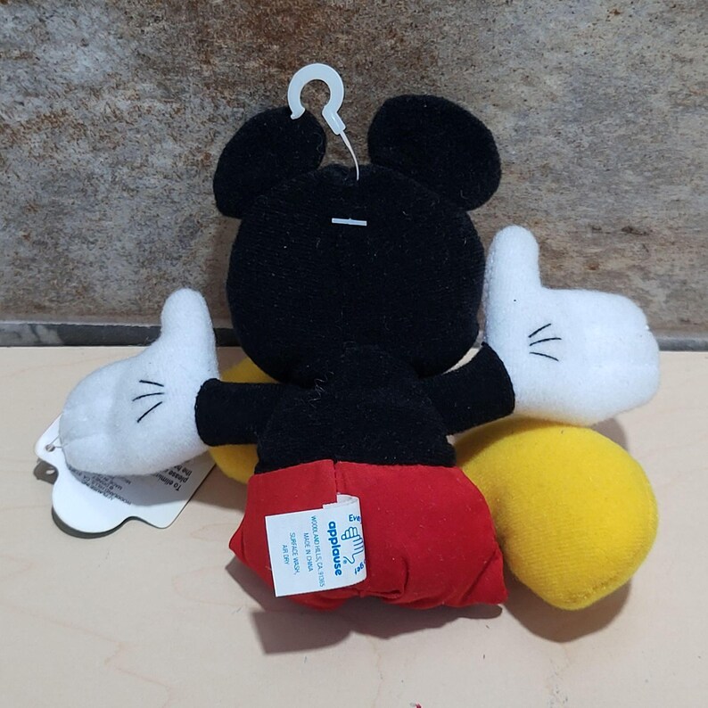 K&ouml;nnte beinhalten: Ein Mickey Mouse Pl&uuml;schtier mit schwarzem Kopf und Ohren, wei&szlig;en Handschuhen, roten Shorts und gelben F&uuml;&szlig;en. Das Spielzeug hat eine wei&szlig;e Plastikschlaufe am Kopf. Ein Etikett ist am Spielzeug befestigt.