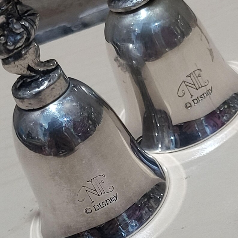 Pu&ograve; includere: Due campane color argento con un logo Disney e le iniziali "NE" incise sul lato. Le campane hanno una superficie lucida e riflettente e una classica forma a campana. Le campane sono probabilmente oggetti decorativi o da collezione.