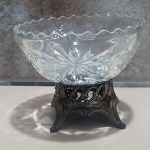 May include: A clear glass bowl with a scalloped edge, set on an ornate, dark bronze-coloured metal stand. The bowl has a starburst pattern on the base. The stand features a decorative, openwork design.