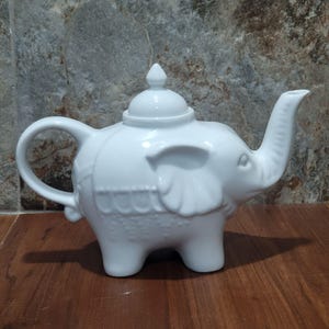 Puede incluir: Una tetera de cerámica blanca con forma de elefante. La tetera tiene un asa curva, un pico en forma de trompa de elefante y una tapa con un pequeño adorno. El cuerpo del elefante está decorado con detalles en relieve.