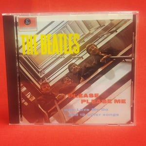 Puede incluir: Una funda de CD con el álbum "Please Please Me" de The Beatles. La portada muestra una foto de los miembros de la banda en una escalera. El título del álbum y el nombre de la banda están en amarillo.