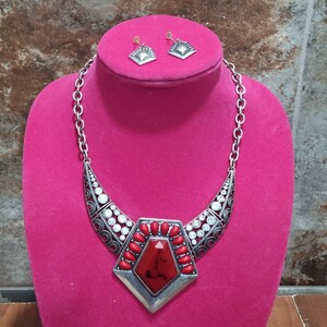 May include: A silver-toned necklace and earring set displayed on a pink bust. The necklace features a large, red, geometric centerpiece with smaller red accents and clear stones. The earrings are small, square, and silver-toned.