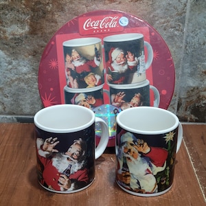 Puede incluir: Conjunto de cuatro tazas de cerámica de la marca Coca-Cola con ilustraciones navideñas vintage de Papá Noel. Las tazas vienen en una caja redonda roja con el logotipo de Coca-Cola.