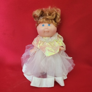 Puede incluir: Una muñeca Cabbage Patch Kid con un vestido de tul blanco y una capa dorada iridiscente. La muñeca tiene el pelo rizado marrón y ojos azules.