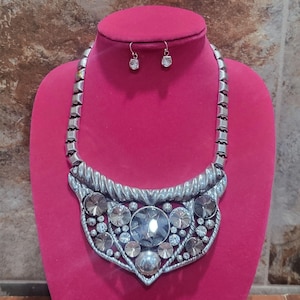 May include: Silver-toned statement necklace and matching earrings. The necklace features a large, central clear crystal surrounded by smaller crystals and a textured metal design. The earrings are small, with a single clear crystal. The jewelry is displayed on a pink mannequin.