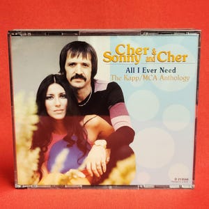 May include: A CD case featuring Cher and Sonny. The album cover has a photo of the duo with the text "Cher & Sonny and Cher" and "All I Ever Need The Kapp/MCA Anthology". The background is a mix of blue, yellow, and green.