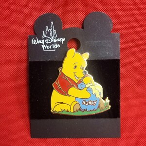 Op de afbeelding: Een gele Winnie de Poeh-speld, met een rood shirt, zittend op groen gras en honing etend uit een blauwe pot met het opschrift "Hunny". De speld is bevestigd aan een zwarte kaart met het "Walt Disney World"-logo.