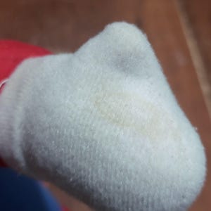 May include: Close-up of a white plush toy hand with a visible stain. The hand is part of a larger toy, with red fabric visible at the wrist. The plush material has a soft, textured appearance.