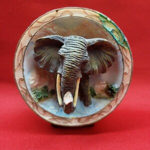 May include: A detailed elephant figurine in a circular frame. The elephant has large ears, tusks, and a textured trunk. The frame has a decorative border. The background shows a water scene with green foliage. The figurine is approximately 10 cm in diameter.
