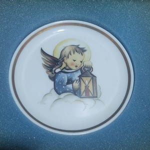 May include: A white porcelain plate with a gold rim features a painting of an angel holding a lantern. The angel is wearing a blue dress with white polka dots and has blonde hair. The angel is sitting on a cloud with a halo above its head.