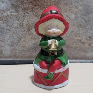 May include: A ceramic Christmas elf figurine. The elf has blonde hair, a red hat with a gold band, and a green outfit with a black belt. The elf is holding a gold bell and sitting on a red drum with white trim. The figurine is approximately 4 inches tall.