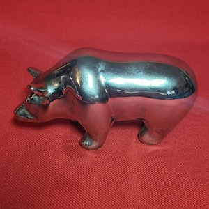 May include: A silver metal sculpture of a bear standing on all fours. The bear is facing to the left and has a shiny, polished finish.