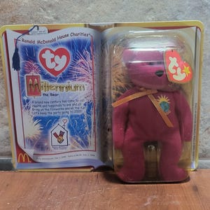 May include: A Millennium the Bear plush toy in a clear plastic case, with a red body and a yellow ribbon. The packaging includes text about the new century and Ronald McDonald House Charities.
