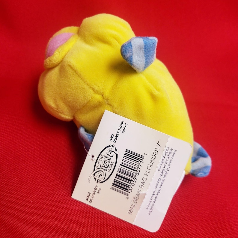 K&ouml;nnte beinhalten: Ein gelbes Pl&uuml;schtier von Fabius aus Arielle, die Meerjungfrau, mit blau-wei&szlig; gestreiften Flossen und einem rosa Kopf. Das Disney Store-Etikett lautet "MINI BEAN BAG FLOUNDER 7" und "MADE EXCLUSIVELY FOR THE DISNEY STORE".
