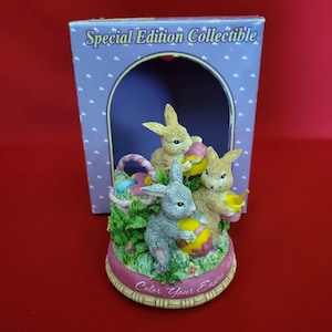 May include: A ceramic musical figurine of three bunnies painting Easter eggs. The bunnies are in a grassy setting with a basket of eggs. The base of the figurine reads "Color Your Easter".