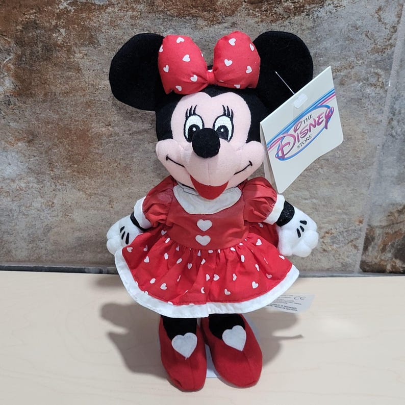 K&ouml;nnte beinhalten: Ein Minnie Mouse Pl&uuml;schtier. Die Puppe hat ein rotes Kleid mit wei&szlig;en Herzakzenten, eine passende Schleife und rote Schuhe. Das Disney Store-Etikett ist angebracht. Das Pl&uuml;schtier ist schwarz, wei&szlig; und rot.