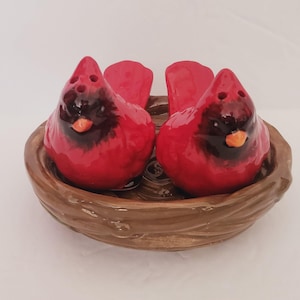 Puede incluir: Un juego de salero y pimentero de cerámica con dos figuras de pájaros cardenales rojos con detalles negros y picos naranjas, anidados en un soporte marrón en forma de nido. Los pájaros tienen agujeros para dispensar. Un artículo de cocina decorativo.