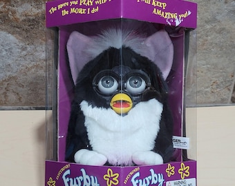 May include: A black and white Furby toy in its original packaging. The electronic toy has large eyes, a yellow beak, and a white chest. The packaging is purple with the word "Furby" and the phrases "The more you PLAY with me the MORE I do!" and "I will KEEP AMAZING you!"