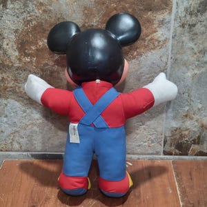 May include: A vintage Mickey Mouse plush doll, viewed from the back. The doll features a black head with large ears, a red shirt, blue overalls with crossed straps, and white gloves. The doll's feet are red with yellow accents.