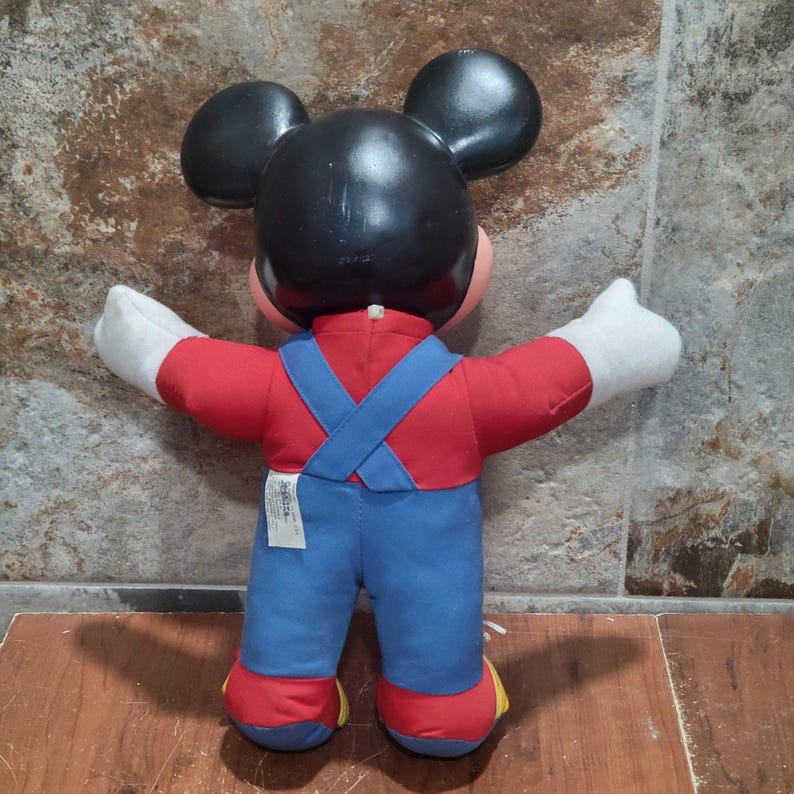 May include: A vintage Mickey Mouse plush doll, viewed from the back. The doll features a black head with large ears, a red shirt, blue overalls with crossed straps, and white gloves. The doll's feet are red with yellow accents.
